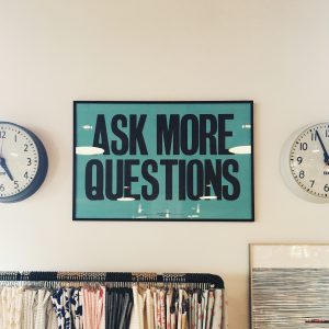 "Ask More Questions" sign on a white wall between two clocks.