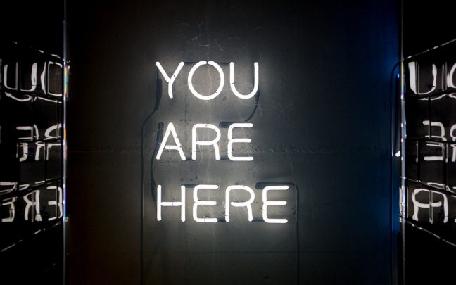 White light neon sign saying "You Are Here" in capital letters on black background.